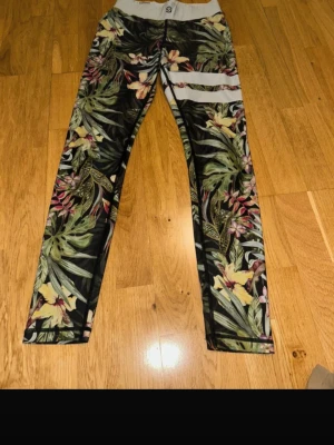 Blommiga träningstights från Stronger - Snygga träningstights från Stronger i storlek small med tropiskt blommönster i grönt, gult, rosa och vitt. Hög midja och vita ränder på ena benet. Tillverkade i stretchigt material av polyester och spandex för maximal komfort.