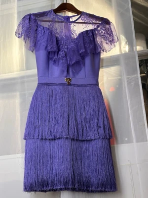 Lila gatsby kortklänning med spets och fransar - Supersnygg lila kortklänning med transparent spets upptill och volangdetaljer över axlarna. Kjolen har två lager långa fransar som ger cool rörelse. Klänningen har rund halsringning och ett dekorativt spänne i midjan. Perfekt för dig som vill sticka ut.