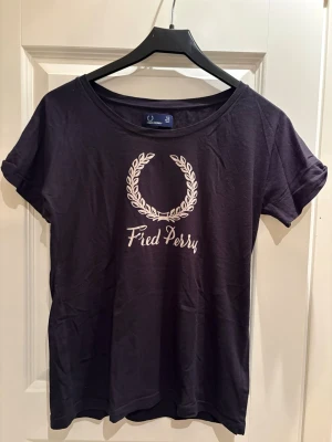 Svart Fred Perry t-shirt med tryck - Svart t-shirt från Fred Perry med klassiskt vitt logotryck framtill. T-shirten har rund halsringning och korta ärmar. Tillverkad i mjuk bomull som är skön att bära. Perfekt för en avslappnad och stilren look.