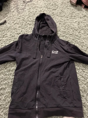 Svart zip-hoodie från EA7 Emporio Armani - Svart hoodie med dragkedja från EA7 Emporio Armani. Tröjan har huva med dragsko, vita logodetaljer på bröstet och ryggen samt fickor framtill. Perfekt för en sportig och avslappnad stil.