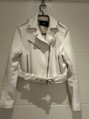 Vit skinnjacka med bälte från Bik Bok - Snygg vit skinnjacka från Bik Bok med croppad passform, silvriga dragkedjor och bälte i midjan. Jackan har axelklaffar och flera fickor med dragkedja för en cool bikerstil. Perfekt för dig som vill sticka ut med en fräsch look.