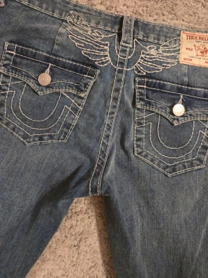 True Religion blå bootcut jeans - Säljer ett par blå jeans från True Religion med ikoniska kontrastsömmar, broderade fickor och snygga silverknappar. Modellen har bootcut och klassisk femficksdesign. Perfekta för dig som gillar detaljer och vill sticka ut. Endast provade alldeles för långa för mig.