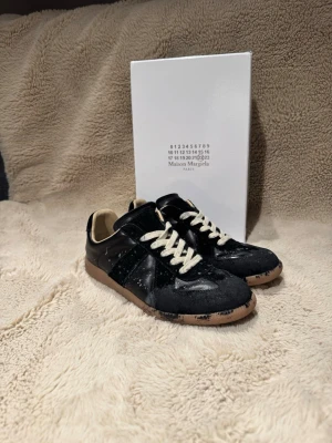 Maison Margiela svarta sneakers - Maison Margiela sneakers i svart skinn och mocka med beige snörning och gummisula. Klassisk låg modell med rund tå och diskreta vita färgstänk på ovandelen och sulan. Snygg kontrast mellan material och detaljer, perfekt för en unik streetwear-look.
