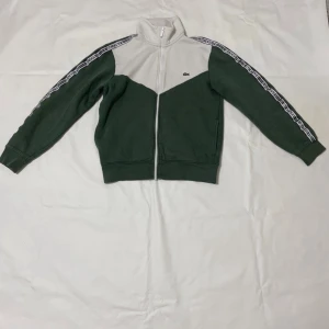 Grön och vit ziptröja från Lacoste - Snygg grön och vit ziptröja från Lacoste med klassisk passform. Tröjan har dragkedja framtill, hög krage och logga på bröstet. Ärmarna har ett mönstrat band längs hela längden. Perfekt för en sportig och avslappnad stil.