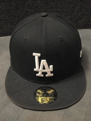 Svart LA Dodgers keps New Era - Säljer en svart New Era 59FIFTY keps med LA Dodgers logga i vitt framtill och ett broderat 'World Series 1981' märke på sidan. Klassisk flat brim och strukturerad passform. Perfekt för dig som gillar streetwear och sportig stil. Priset är ej hugget i sten. Vid frågor hör av er