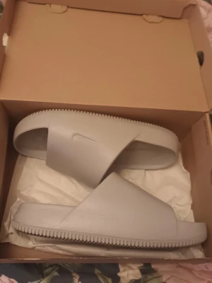 Gråa slides från Nike - Säljer ett par stilrena gråa slides från Nike. Tofflorna har en bred rem över foten med den klassiska Nike-loggan och ribbad sula för extra grepp. Perfekta för chill dagar hemma eller till stranden. Tillverkade i syntetmaterial för en lätt och bekväm känsla. I nyskick endast använda 1 timme ungefär finns på eskilsvägen vid Haninge centrum men kan träffas upp i närheten eller skicka på posten swish eller kontanter 