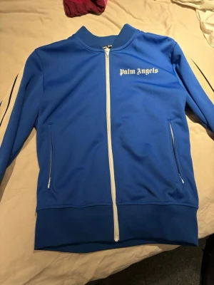 Blå ziptröja från Palm Angels - Snygg blå ziptröja från Palm Angels med vita detaljer och dragkedja framtill. Tröjan har vita ränder längs ärmarna och två fickor med dragkedja. Perfekt för en sportig och trendig look.