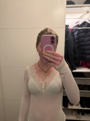 Vit långärmad topp med spets - Säljer en vit långärmad topp med v-ringning och fina spetsdetaljer längs halsringningen och ärmsluten. Toppens material är tunt och stretchigt, vilket ger en tight passform. Perfekt att styla med jeans eller kjol.