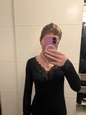 Svart långärmad spetsblus - Snygg svart långärmad blus med v-ringning och spetsdetaljer längs halsringningen. Toppens material är tunt och stretchigt, vilket ger en tight passform. Perfekt för dig som gillar en stilren och feminin look.