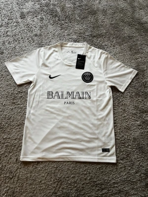 PSG x Balmain jersey - Snygg vit t-shirt från Nike i samarbete med PSG och Balmain. Tröjan har Dri-FIT-material som andas, rund halsringning och tryckta loggor från Nike, PSG och Balmain Paris på bröstet. Perfekt för träning eller streetwear.