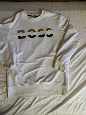 Vit BOSS sweatshirt med tryck - Vit sweatshirt från BOSS med stort tryck i svart, brunt och vitt på bröstet. Tröjan har rund hals, ribbade muddar och avslut. Perfekt för en avslappnad och stilren look.