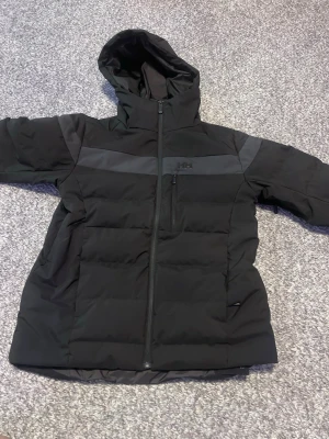 Svart pufferjacka från Helly Hansen  - Säljer en svart pufferjacka från Helly Hansen med huva och hel dragkedja. Jackan har en diskret logga på bröstet, breda quiltade paneler och en mörkgrå rand över bröst och rygg. Insidan har meshficka och RECCO-reflektor. Perfekt för kalla dagar. Jag tog på mig den i några veckor bara! 
