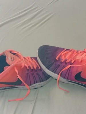 Nike Free sneakers i rosa och lila - Nike Free sneakers med mesh-ovandel i en snygg gradient från rosa till lila och svarta detaljer. Skorna har en vit och svart sula med flexibelt mönster och klassisk Nike-logga på sidan. Utrustade med snörning och platt sula, perfekta för träning eller vardagsstil.