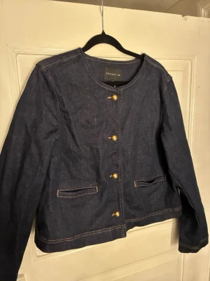 Mörkblå jeansjacka från STOCKH LM - Säljer en stilren mörkblå jeansjacka från STOCKH LM i klassisk modell. Jackan har rund halsringning, guldiga knappar framtill och två fickor. Snygga kontrastsömmar och lång ärm ger en clean look. Perfekt till jeans eller kjol.