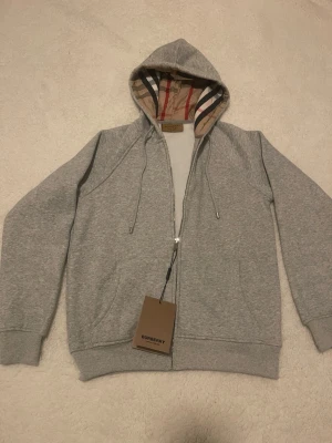 Grå hoodie från Burberry med dragkedja - Snygg grå hoodie från Burberry med dragkedja och klassiskt rutigt foder i huvan. Hoodien har två fickor fram, dragsko i huvan och diskret Burberry-logga broderad på bröstet. Tillverkad i mjuk bomullsblandning för skön känsla.