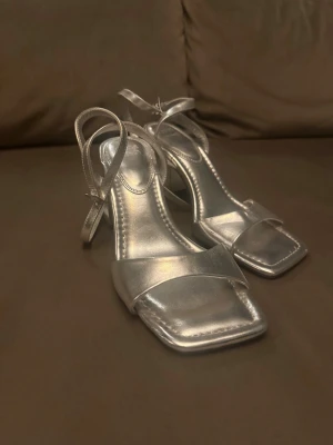 Silvriga sandaler med klack och remmar - Säljer ett par stilrena silvriga sandaler med fyrkantig tå, bred rem över foten och justerbara ankelremmar. Skorna har en stabil klack och platt sula framtill. Perfekta för dig som vill sticka ut med metallic-look.