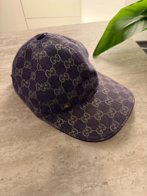 Blå keps - Snygg blå keps i denim-look med klassiskt monogram över hela kepsen. Justerbar rem bak och insida i ljus bomull. Perfekt för dig som vill ha en exklusiv och trendig accessoar med tydlig logga.