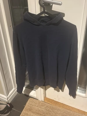 Mörkblå hoodie med huva - Är 163cm. Inga hål. En enkel och stilren mörkblå hoodie med huva. Hoodien har långa ärmar och ribbade muddar vid ärmslut och nederkant. Perfekt för en avslappnad look och passar till det mesta. Materialet känns mjukt och bekvämt mot huden.
