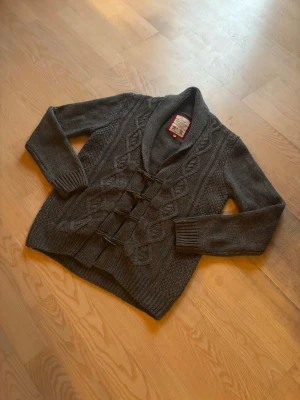 UNIK CARDIGAN - Snygg mörkgrå stickad kofta med grov kabelstickning och fyra dekorativa träknappar framtill. Koftan har bred krage och långa ärmar med ribbade muddar. Perfekt för kyliga dagar och enkel att matcha med jeans eller chinos. Passar bra om man är runt 180 beroende på vikt! PRIS GÅR ATT DIKSUTERA