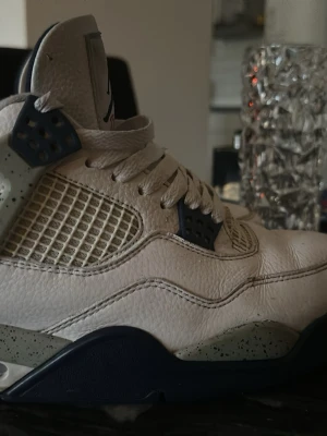 Nike Air Jordan 4 vit/blå sneakers - Säljer ett par Nike Air Jordan 4 sneakers i vitt skinn med blå och grå detaljer. Skorna har meshpaneler på sidorna, snörning och en robust sula med stänkdetaljer. Klassisk Jumpman-logga på plösen och cool retrostil.