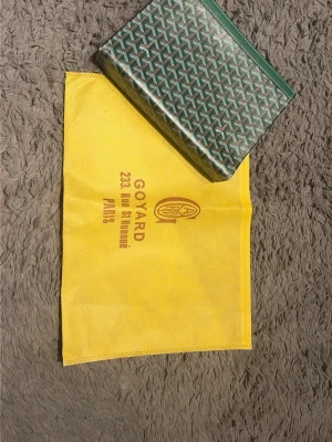 Grön kuvertväska! - Snygg grön kuvertväska. med det klassiska vita och gråa mönstret. Väskan har en rektangulär form och är tillverkad i skinn med detaljer i grönt. Perfekt accessoar för dig som vill sticka ut med något exklusivt.