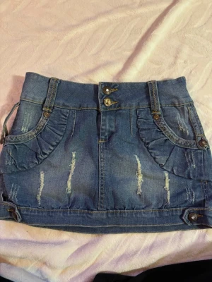  jeanskjol - Säljer en blå kort jeanskjol med massa fina detaljer och volang vid fickorna. Kjolen har dubbla knappar framtill, dekorativa sömmar och bälteshällor. Insidan är fodrad med blommigt tyg för extra stilpoäng. Osäker på storlek skulle gissa på S