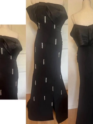 Svart axelbandslös maxiklänning / balklänning  - Elegant svart axelbandslös maxiklänning med stor volang upptill och dekorativa pärl- och paljettbroderier över hela klänningen. Figurnära passform med slits framtill. Perfekt för dig som vill sticka ut på nästa event.