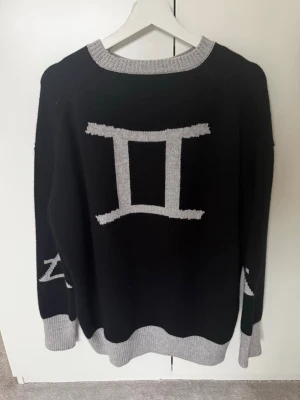 Cloeys cashmere stickad tröja med svart zodiakmönster - Cool svart stickad tröja med grått tvillingarna-symbol på bröstet och gråa detaljer vid hals, ärmslut och nederkant. Oversized. Använd 3 ggr! Så som NY!!