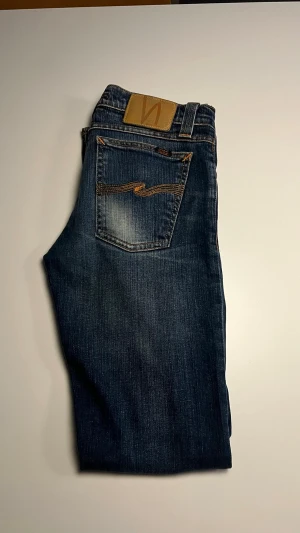 Mörkblå jeans från Nudie Jeans - Snygga mörkblå jeans från Nudie Jeans med klassisk femficksmodell och kontrastsömmar. Jeansen har en rak passform och är tillverkade i slitstark bomull med tydliga Nudie-detaljer på bakfickan och läderpatch i midjan.