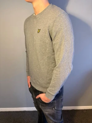 Lyle & Scott tröja - Klassisk grå sweatshirt från Lyle & Scott med rund halsringning och den ikoniska gula fågel-loggan broderad på bröstet. Tröjan har ribbade muddar vid ärmslut och nederkant, perfekt för en avslappnad och stilren look. Storlek M. Modellen är 184cm.