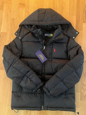 Svart dunjacka från Polo Ralph Lauren - Snygg svart dunjacka från Polo Ralph Lauren med huva och dragkedja framtill. Jackan har en quiltad design, bröstficka med dragkedja och den klassiska röda loggan broderad på bröstet. Perfekt för kalla dagar och riktigt stilren.