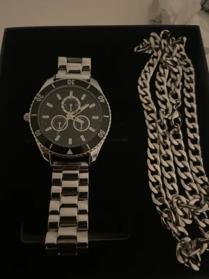 Stilren silverfärgad klocka plus silverkedja från finmärkt Pier One - Snygg klocka i silverfärgad metall med svart urtavla och vita markeringar. Klockan har länkarmband och flera små urtavlor för en sportig look. Perfekt accessoar för dig som gillar klassisk och modern stil. Kedja i matchande silverfärg medföljer.