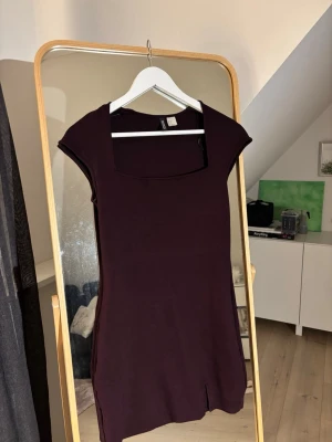 Mörklila kort klänning med slits - Snygg mörklila kort klänning med fyrkantig ringning och liten slits framtill. Klänningen har korta ärmar och en figurnära passform som framhäver siluetten. Perfekt för dig som gillar minimalistisk och stilren design.