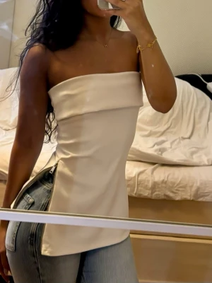 Vit axelbandslös tubtopp med dragkedja - Säljer en vit axelbandslös tubtopp  med bred kant upptill och cool dragkedjedetalj längs sidan. Toppen har en clean och stilren look som passar perfekt till jeans eller kjol.