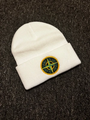 Vit mössa från Stone Island - Snygg vit mössa från Stone Island med klassisk uppvikt kant och det ikoniska broderade logomärket framtill i grönt och gult. Tillverkad i mjukt stickat material som håller dig varm och stilren under kalla dagar.