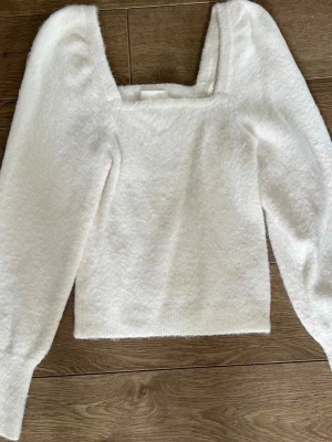 Vit fluffig stickad tröja H&M - Supersöt vit stickad tröja från H&M med fluffig känsla och fyrkantig ringning. Tröjan har långa ärmar och sitter snyggt med en lite croppad passform. Perfekt till både jeans och kjol för en trendig look.