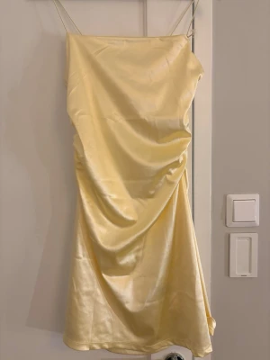 Ljusgul satin klänning med smala band - Säljer en ljusgul kort klänning i satin med smala axelband och draperad front. Klänningen har en glansig finish och en enkel, elegant design som känns både trendig och stilren. Perfekt för dig som vill sticka ut med en mjuk pastellfärg.