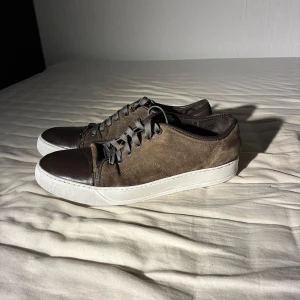 Bruna/gråa mockasneakers med skinn-detaljer - Snygga sneakers i brun/grå mocka med mörkbruna skinnpartier på tån och hälen. Skorna har vita, platta sulor och grå snörning, man får även till blå snören. Perfekta för dig som gillar en stilren och klassisk look med en twist av olika material. OBS litet hål i innersulan men den går att byta och lite slitet i hälen men okej skick.