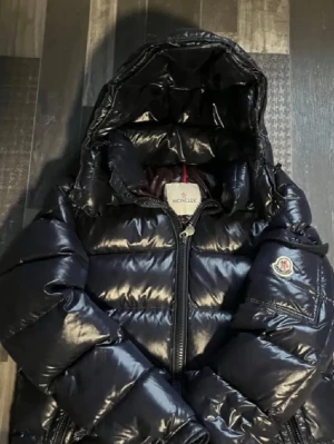 Moncler Maya  - Säljer en svart, glansig pufferjacka från Moncler med huva och dragkedja framtill. Jackan har Moncler-logga på ärmen och är riktigt fluffig och varm, perfekt för kalla dagar. Materialet är syntet och dun, vilket ger en schysst look och maxad komfort.