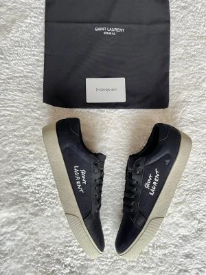 Svarta Saint Laurent sneakers - Svarta sneakers från Saint Laurent med vit text på sidan och guldfärgad logga på plösen. Skorna har snörning, rund tå och en beige platt sula. Tillverkade i canvas och skinn för en stilren och modern look.