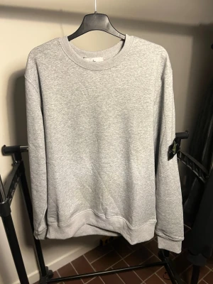  Stone Island - Snygg grå sweatshirt från Stone Island med klassisk rund halsringning och ribbade muddar. Tröjan har den ikoniska Stone Island-loggan på ärmen och är tillverkad i mjukt bomullsmaterial. Perfekt för en clean och avslappnad stil.