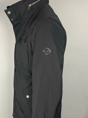 Svart vindjacka från J.Lindeberg - Stilren svart vindjacka från jlindeberg med hög krage, knapp- och dragkedjestängning samt flera fickor framtill. Jackan har en diskret logga på ärmen och är tillverkad i slitstark polyester. Perfekt för dig som vill ha en snygg och funktionell jacka.