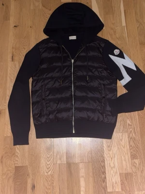 Moncler cardigan - Snygg svart cardigan från Moncler med quiltad kropp, stickade ärmar och huva. Dragkedja framtill och stor vit zigzagdetalj på ena ärmen med Moncler-logga. Perfekt för dig som vill ha en trendig och bekväm jacka.