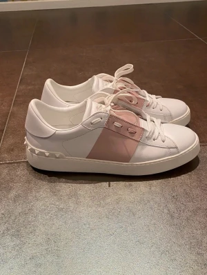 Valentino Garavani sneakers vit/rosa - Snygga Valentino Garavani sneakers i vitt skinn med en bred rosa panel och matchande rosa detaljer vid snörningen. Skorna har klassisk snörning och coola nitar på hälen för extra edge. Perfekta för dig som vill ha en stilren men unik look.