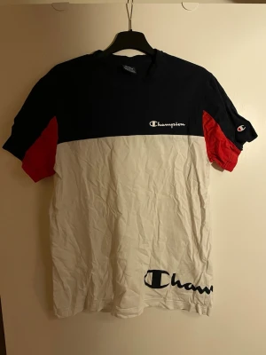 Champion t-shirt i färgblock M - Snygg t-shirt från Champion med färgblock i marinblått, vitt och röda detaljer på ärmarna. Klassisk logga på bröstet och ärmen samt stor logga nertill. Skön passform och tillverkad i mjuk bomull.