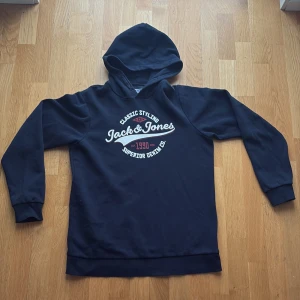 Mörkblå hoodie från Jack & Jones - Snygg mörkblå hoodie från Jack & Jones med vit och röd logga på bröstet. Klassisk huva och långa ärmar. Tillverkad i mjukt material som känns skönt mot huden. Perfekt för en avslappnad och cool stil.