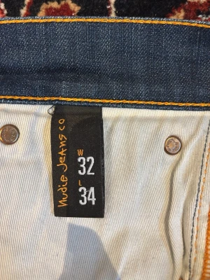 Blå jeans från Nudie Jeans Co W32 L34 - Snygga blå jeans från Nudie Jeans Co i modellen W32 L34. Tillverkade av 100% ekologisk bomull med klassiska orange sömmar och detaljer i metall. Perfekta för dig som gillar stilrena och hållbara jeans med schysst passform.
