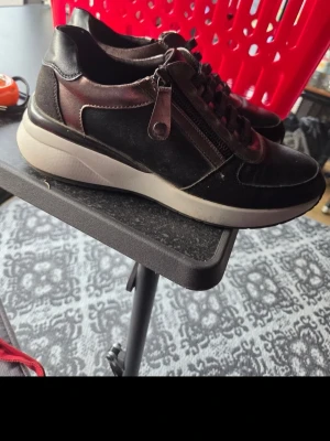 Svarta sneakers med dragkedja - Snygga svarta sneakers med vit platt sula och dragkedja på sidan. Skorna har rund tå och snörning framtill. Materialet ser ut att vara skinn eller syntet, vilket ger en stilren look.