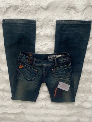 Mörkblå jeans från Freesoul, stl 26 - Snygga mörkblå jeans med låg midja och utsvängda ben från Freesoul med coola detaljer som dubbla knappar, kontrastsömmar och unika fickor framtill. Jeansen har en klassisk femficksdesign och orange logotypdetalj på ena fickan. Passar en xs 🩷