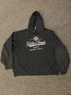Svart Golden Nugget hoodie - Snygg svart hoodie med vit text och tryck på bröstet där det står 'Golden Nugget Original Las Vegas Nevada'. Klassisk känguruficka framtill och stor huva. Perfekt för dig som gillar streetwear och vill ha en bekväm och stilren look.  Passar mig bra och jag är 185cm